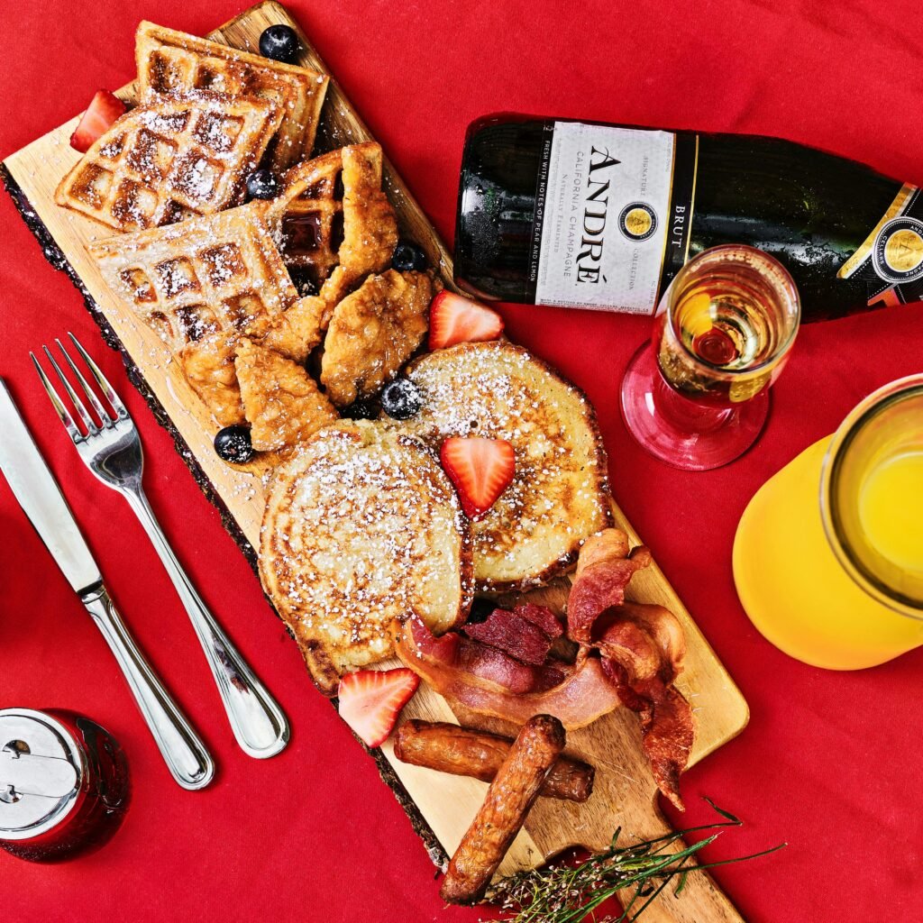 Top view of a brunch platter with waffles, pancakes, bacon, and champagne on a red tablecloth.