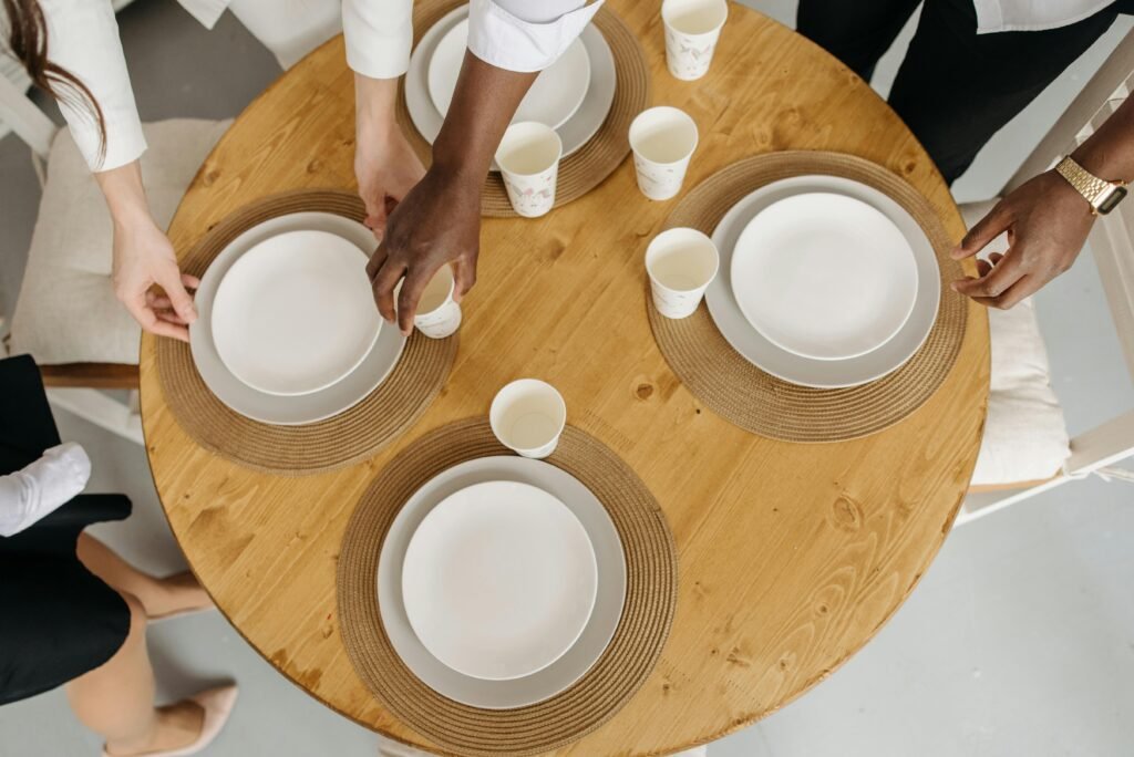 Three adults setting a round wooden table with ceramic plates and paper cups indoors.