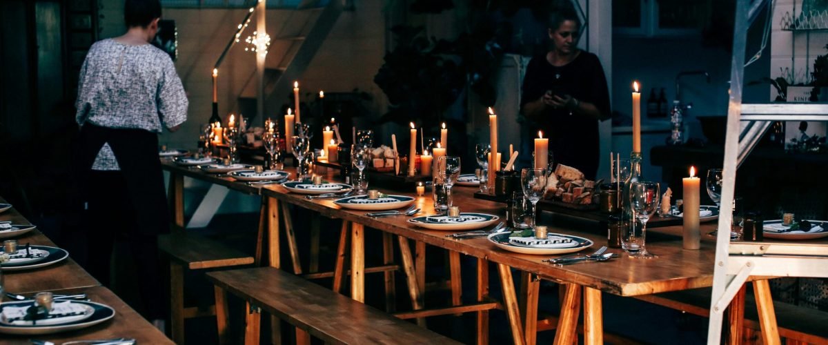 Anonymous women serving wooden tables with plates and glassware among burning candles while preparing for banquet in modern place