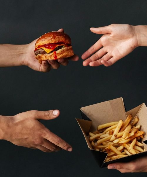 Close-up of hands exchanging a burger and fries against a dark background.